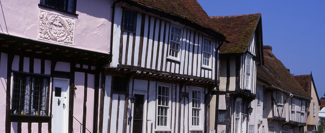 A row of crooked timber-framed medieval houses, one painted Suffolk pink, on a sunny village street